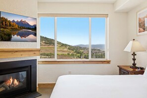 Een hotelkamer met een bed, een open haard, een groot raam met uitzicht op een bergachtig landschap en een lamp op een nachtkastje.