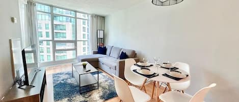 A modern living room with a sofa, dining table, and chairs, a flat-screen TV, and a large window with curtains.