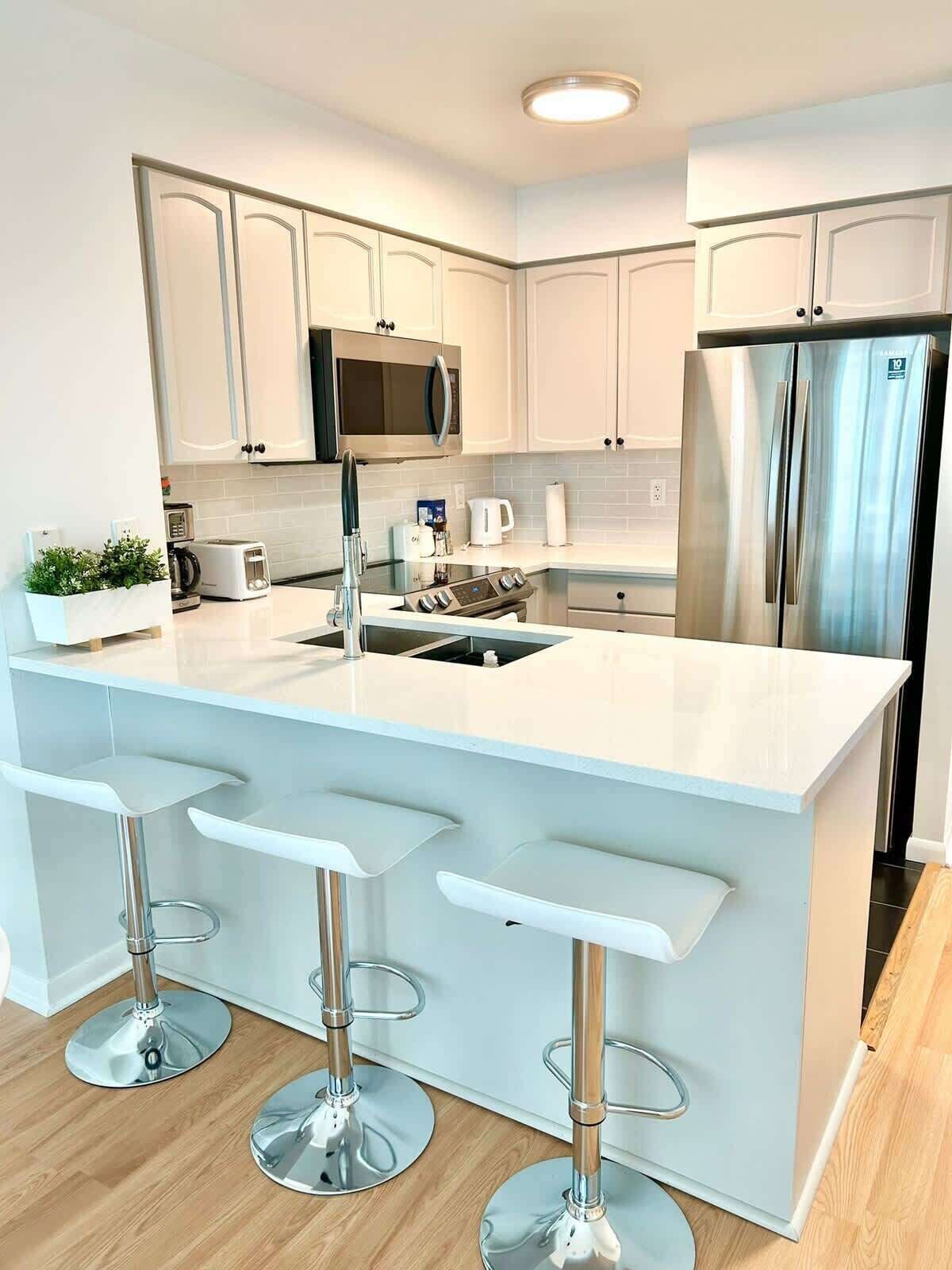 Una cocina moderna con armarios blancos, un refrigerador de acero inoxidable, un microondas y una cafetera. Hay tres taburetes de bar con bases cromadas.