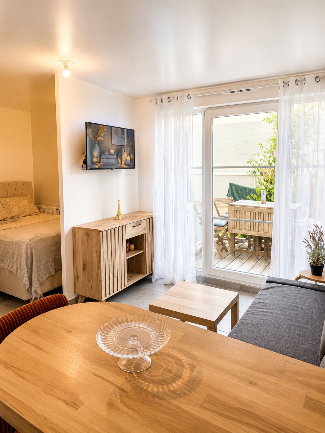 A room with a bed, a wooden cabinet, a dining table with a glass cake stand, and a balcony with a table and chairs.