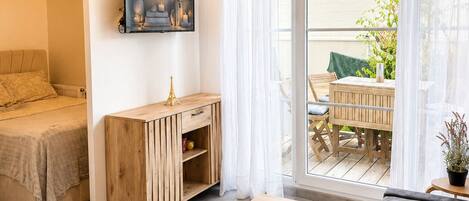 A room with a bed, a wooden cabinet, a dining table with a glass cake stand, and a balcony with a table and chairs.