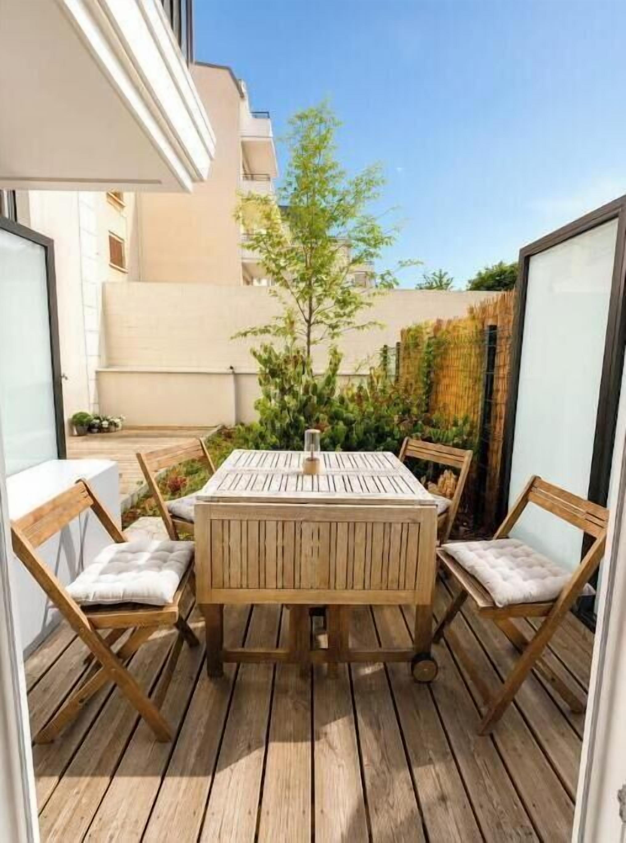 An outdoor wooden deck with a round table and four chairs, surrounded by a building with a white exterior and a clear sky.