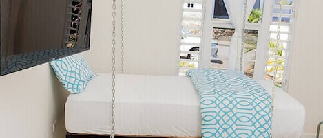 A room with a bed suspended from the ceiling, a flat-screen TV mounted on the wall, and a view of the outside through a window.