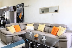 A living room with a sectional sofa, a glass coffee table, and a kitchen area with appliances. - Beyond Escape-solar-powered-3bdrm-near Ocho Rios (Boscobel)