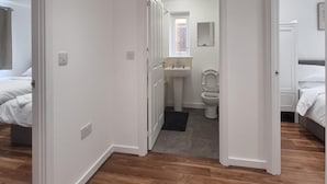 A hotel room with a wooden floor, a bed, a bathroom with a sink and toilet, and a hallway leading to another room.