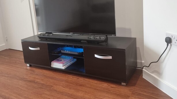 A flat-screen TV on a black entertainment unit with two drawers, a power outlet, and a door in the background.