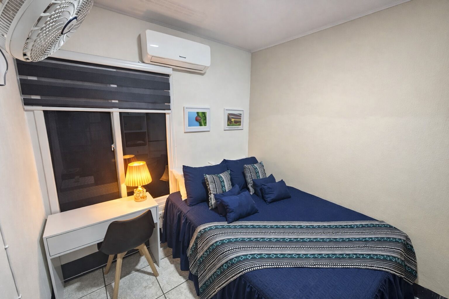 Un dormitorio con cama, escritorio, silla, aire acondicionado y ventana con persianas.