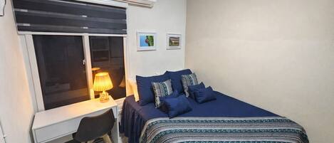 Un dormitorio con cama, escritorio, silla, aire acondicionado y ventana con persianas.