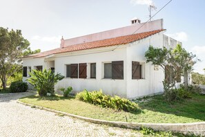 A single-story building with a tiled roof, a flat facade, and a small garden area with plants and a paved driveway. - Vrbo Property (Tavira)