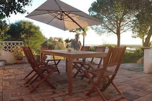 An outdoor patio with a table and chairs, a large umbrella, and a view of the sea. - Cabanas Jardim da Ria: a private home with a large garden, a pool, and views of the estuary (Tavira)