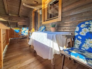 Eine Holzhütte mit Essbereich, ausgestattet mit einem Tisch, einer weißen Tischdecke, einer Topfpflanze und stuhlförmigen Sitzgelegenheiten mit Blumenmuster.
