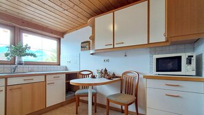 A kitchen with wooden cabinets, a white countertop, a dining table with chairs, a microwave, and a window with a view of greenery. - Pessenteiner by Interhome (Saalfelden am Steinernen Meer)