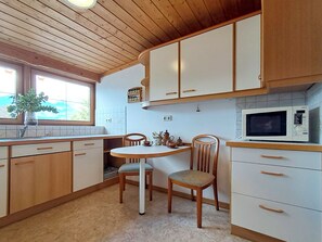 A kitchen with wooden cabinets, a white countertop, a dining table with chairs, a microwave, and a window with a view of greenery. - Pessenteiner by Interhome (Saalfelden am Steinernen Meer)