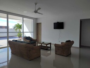 A spacious room with a large window, a ceiling fan, a flat-screen TV mounted on the wall, and wicker furniture.