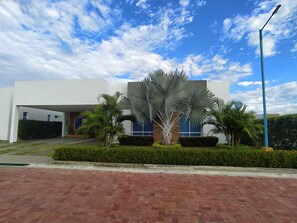 A modern building with a white facade, a covered entrance, and a landscaped area with palm trees and hedges.