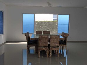 A room with a glass table and wicker chairs, large windows, and a ceiling fan.