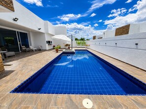 A rectangular swimming pool with blue tiles, surrounded by a patio with outdoor furniture and potted plants.