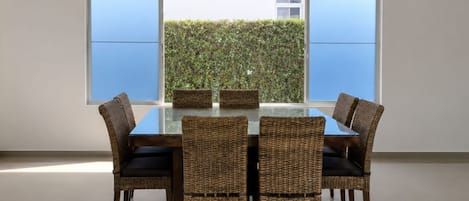 A dining area with a glass table surrounded by wicker chairs, large windows, and a view of greenery outside.