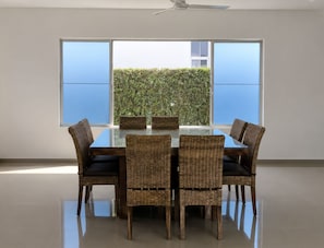 A dining area with a glass table surrounded by wicker chairs, large windows, and a view of greenery outside.