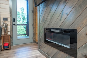 Una habitación con pared de madera, chimenea de vidrio, una puerta con vistas y una pequeña mesa de madera con un jarrón.