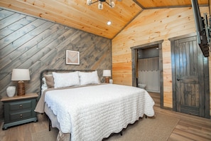Un dormitorio con techo de madera, una cama con ropa de cama blanca, una mesita de noche con lámpara y una puerta.