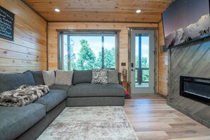 A modern living room with a sectional sofa, a fireplace, and a large window offering a view of the outdoors. - Pigeon Forge Sunsets- Cabin With Mountain Views (Sevierville)