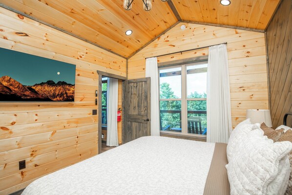 A wooden cabin bedroom with a bed, a window with curtains, a painting of mountains, and a ceiling light fixture. - Pigeon Forge Sunsets- Cabin With Mountain Views (Sevierville)