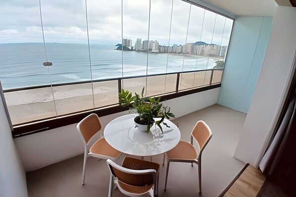 Varanda com mesa redonda e cadeiras, um vaso de planta e vista para o mar e prédios.