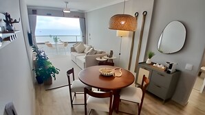 A modern living room with a round wooden table, two chairs, a sofa, and a small side table. There is a large window with a view of the sea, a hanging light fixture, and a mirror on the wall. - SHB - Alto Padrão com Vista  (Guarujá)