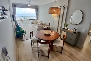 A modern living room with a round wooden table, two chairs, a sofa, and a small side table. There is a large window with a view of the sea, a hanging light fixture, and a mirror on the wall. - SHB - Alto Padrão com Vista  (Guarujá)