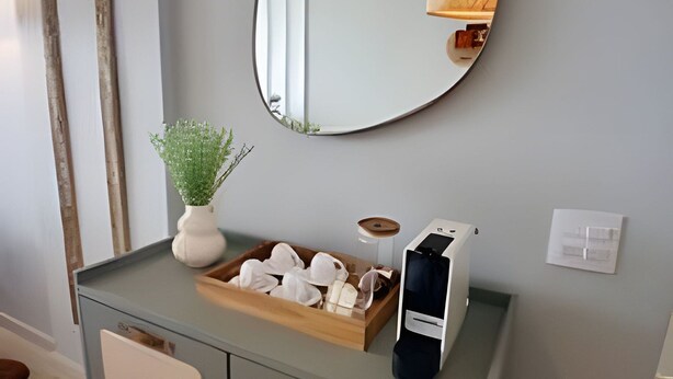 Una kitchenette pequeña con un espejo, una planta, una bandeja de tazas blancas y una cafetera.
