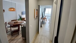 A modern hallway with a dining area featuring a wooden table and chairs, a hanging light fixture, and a view into a living space with a balcony. - SHB - Alto Padrão com Vista  (Guarujá)