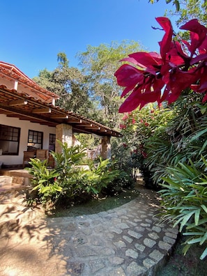 A stone pathway leading to a building with a tiled roof and a garden with various plants and a red flower.