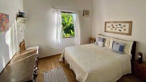 Un dormitorio con cama, ventana con cortinas, mesita de noche, estante en la pared y un cuadro enmarcado.