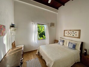 Ein Schlafzimmer mit Bett, einem Fenster mit Vorhängen, einem Nachttisch, einem an der Wand montierten Regal und einem gerahmten Bild.
