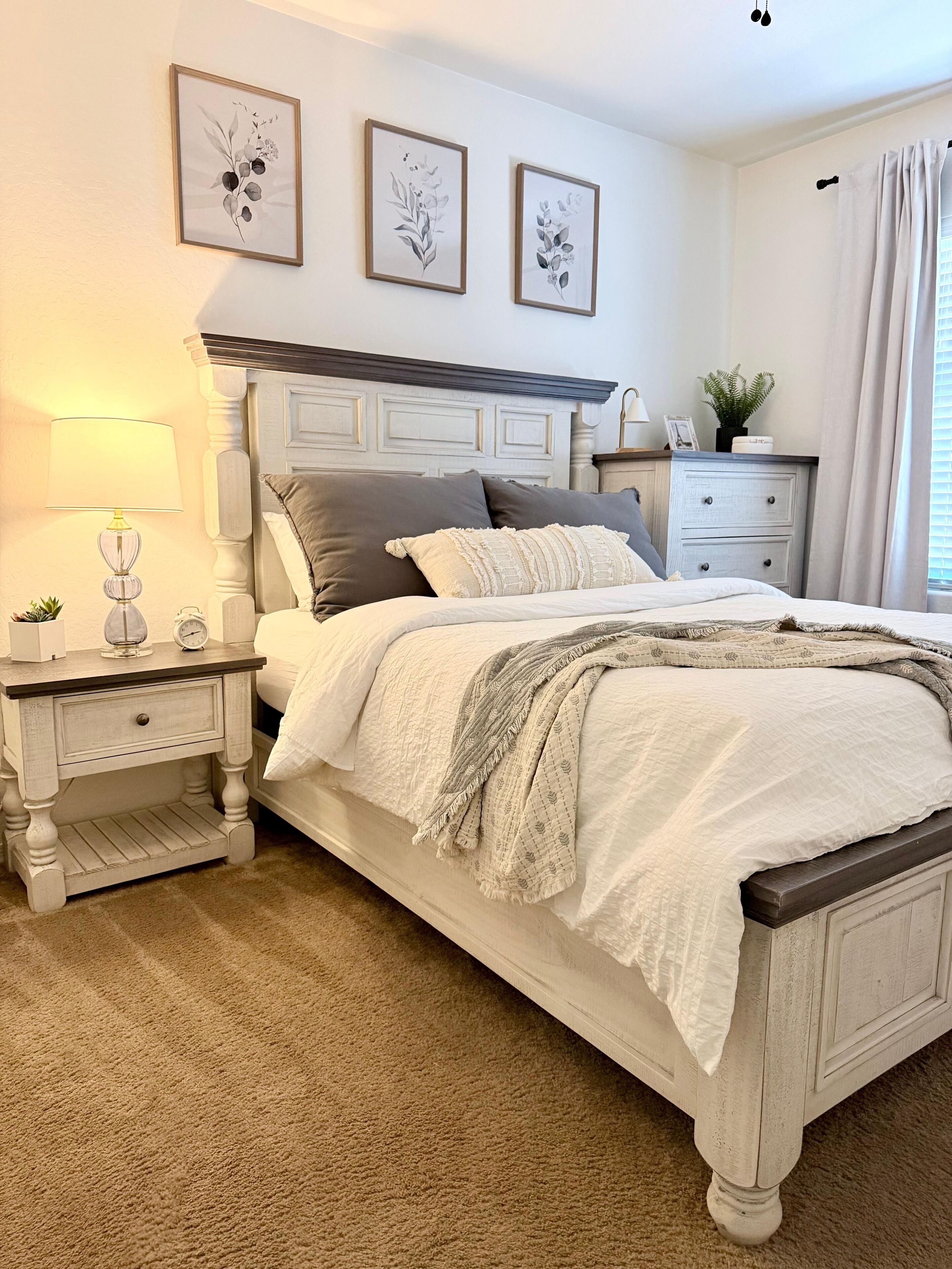 A bedroom with a bed, nightstands, a lamp, framed artwork, and a window with curtains.