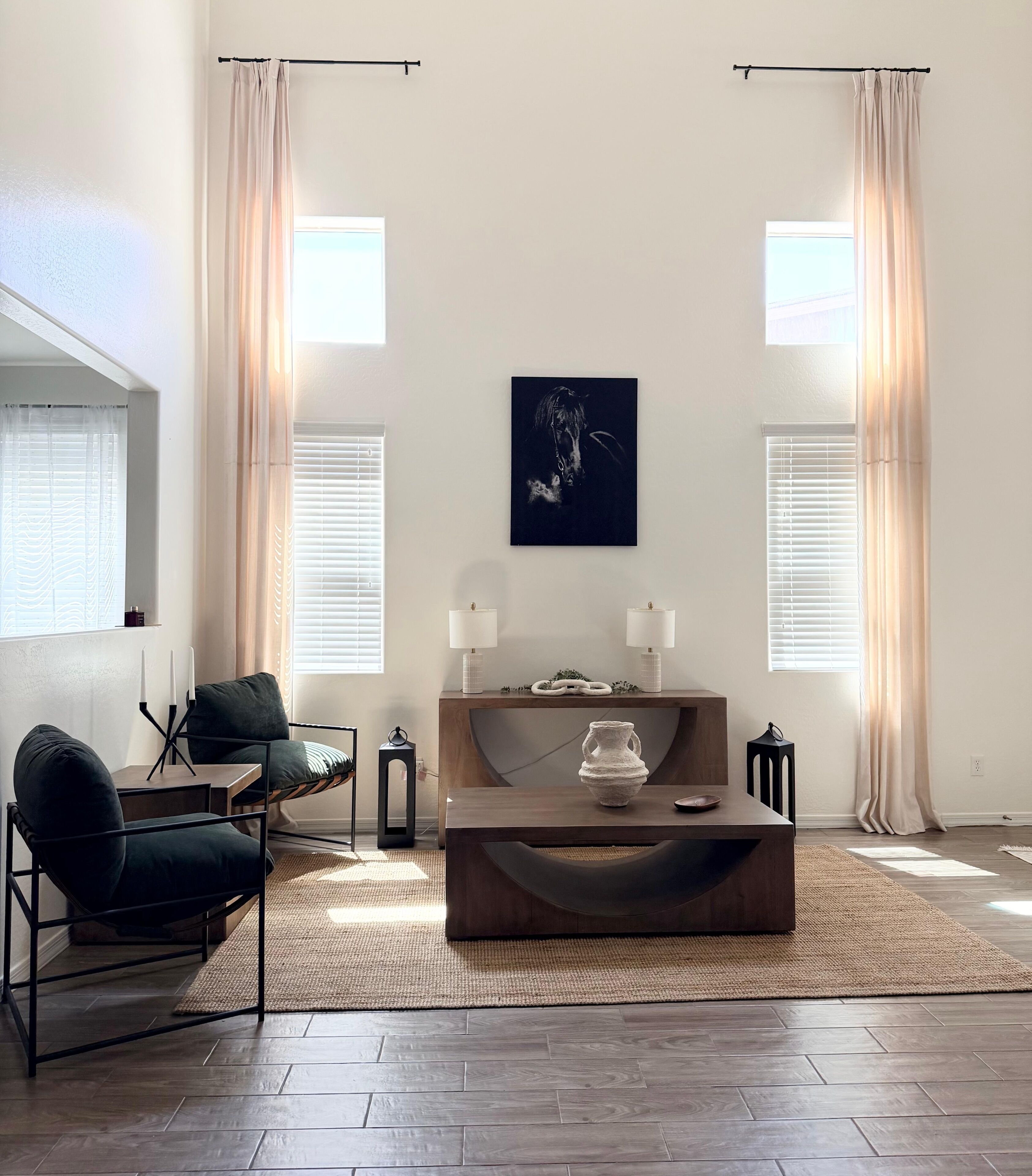 A modern living room with a dark wood coffee table, a green armchair, and a dark sofa. There are two white lamps on a wooden console table, and a painting hangs on the wall.