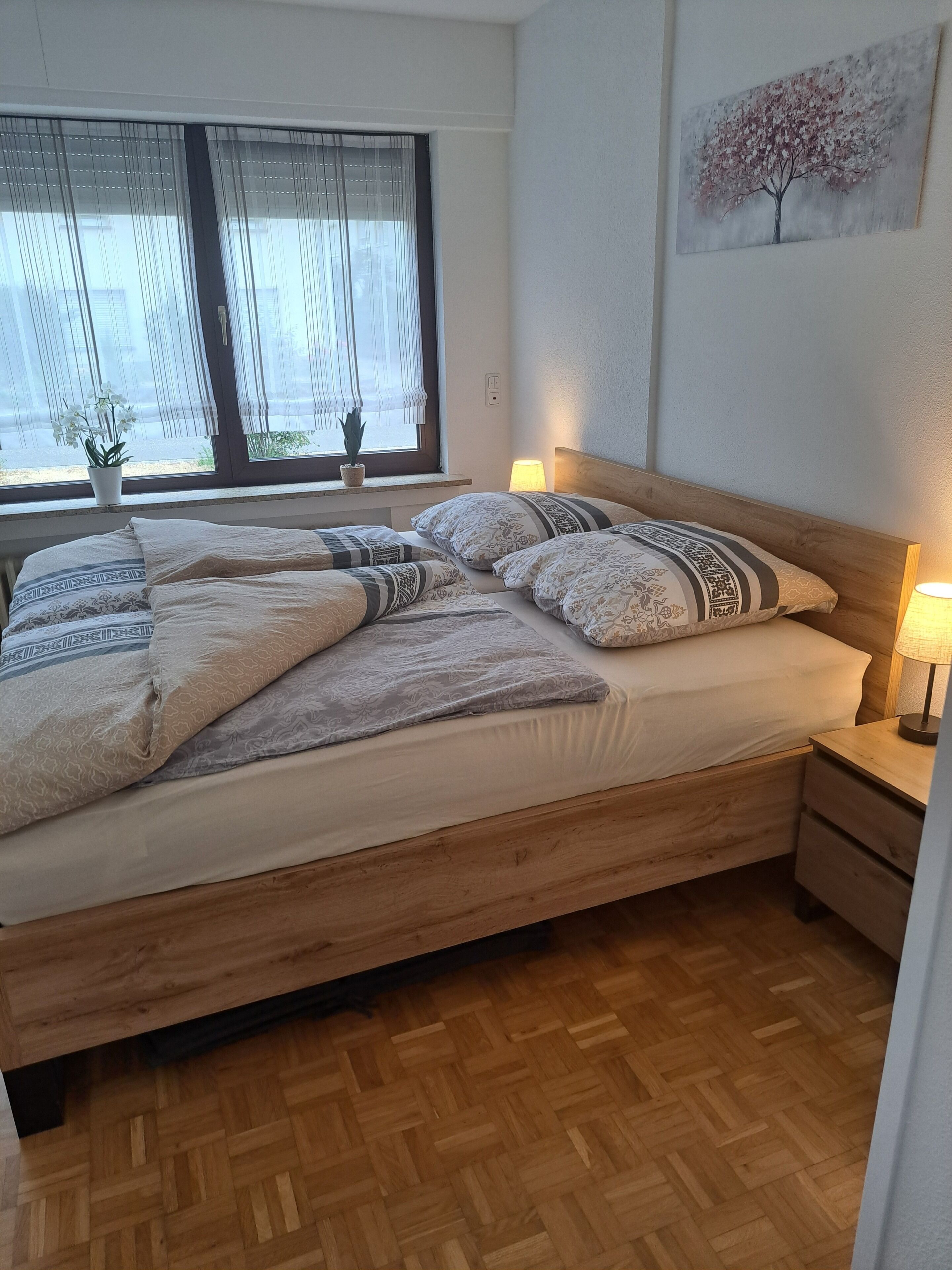 A neatly made bed with patterned bedding, a bedside lamp, a wooden nightstand, and a window with sheer curtains.