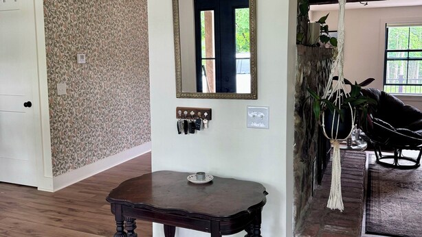 A room with a wooden floor, a dark wooden table, a wall-mounted mirror, a door, a wall with a leaf pattern, a hanging macrame plant, and a staircase.