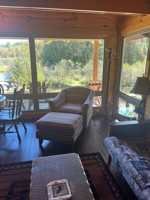 A wooden interior with a view of a natural landscape, a wooden bench, a cushioned chair, and a table. - Serene 2-bedroom cabin on Little Manistee River -Fisherman's paradise (Manistee)