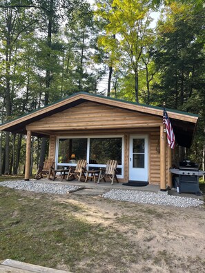 A wooden cabin with a porch, outdoor seating, and an American flag. - Serene 2-bedroom cabin on Little Manistee River -Fisherman's paradise (Manistee)