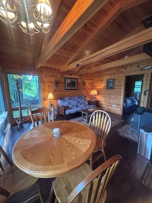Interior de una cabaña de madera con una mesa redonda para comer, sillas, un sofá y una lámpara.