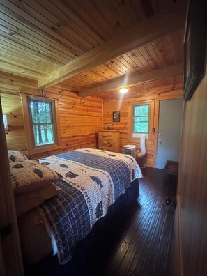 Interior de una cabaña con techo de madera, dos camas con colchas, un aparador y una ventana con vista al bosque.
