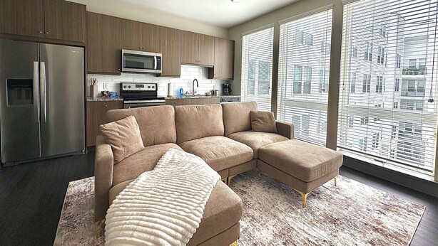 A modern kitchen with stainless steel appliances, a beige sectional sofa, and large windows with blinds. - Luxury Studio Apartment - 10 mins from Downtown (Houston)