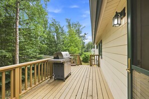 A wooden deck with a barbecue grill, a railing, and a house in the background. - Spacious Lakeside Home with Firepit, Wraparound Deck, Kayaks & Gym (Anchorage)