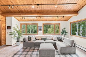 A modern living room with a large sectional sofa, a small ottoman, and a plaid rug. There are three large windows providing views of the forest. - Spacious Lakeside Home with Firepit, Wraparound Deck, Kayaks & Gym (Anchorage)