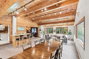 A spacious room with a wooden ceiling, a dining table with chairs, a sofa, and a large window offering a view of trees. - Spacious Lakeside Home with Firepit, Wraparound Deck, Kayaks & Gym (Anchorage)