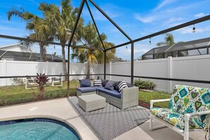 A covered outdoor seating area with a pool, white fence, and palm trees.