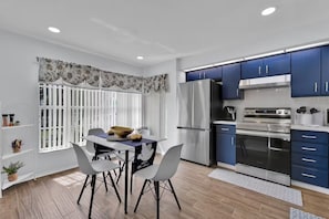 A modern kitchen with stainless steel appliances, a dining area with a table and chairs, and a window with vertical blinds.