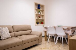 A beige sofa with a patterned pillow, a dining table with a lace tablecloth, white chairs, and a wall-mounted shelf with decorative items. - Experience Palermo in complete comfort: a bright, cozy, and centrally located home. (Palermo)